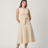 A woman modeling a sleeveless, knee-length beige dress with a matching waist tie, paired with brown ankle boots, against a light gray studio background.