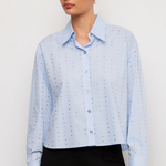 A woman wearing a light blue long-sleeve button-up shirt with a subtle dotted eyelet pattern, paired with black pants, against a plain light gray studio background.