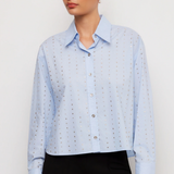 A woman wearing a light blue long-sleeve button-up shirt with a subtle dotted eyelet pattern, paired with black pants, against a plain light gray studio background.