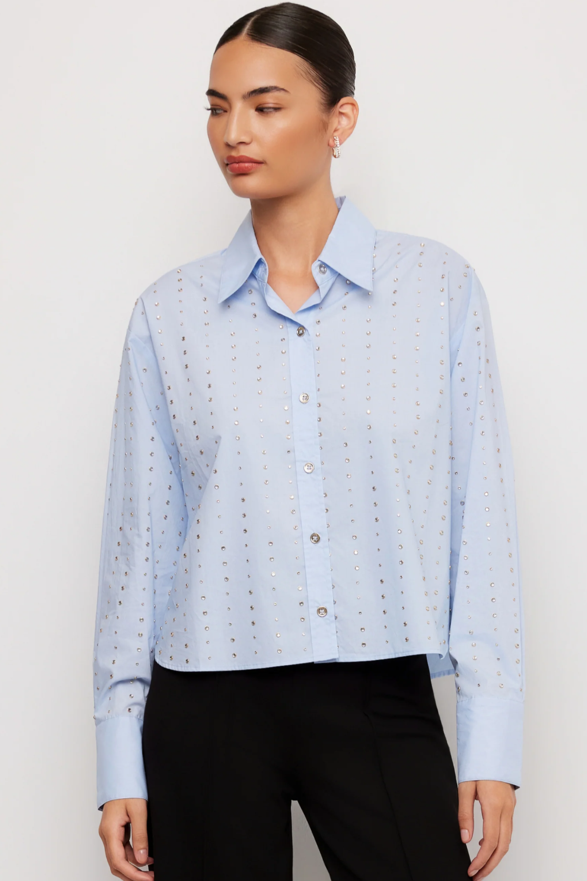 A woman wearing a light blue long-sleeve button-up shirt with a subtle dotted eyelet pattern, paired with black pants, against a plain light gray studio background.