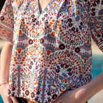 A close-up of a woman wearing a short-sleeve V-neck blouse with a colorful bohemian floral and paisley print in orange, blue, black, and white, paired with dark blue jeans.