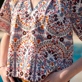 A close-up of a woman wearing a short-sleeve V-neck blouse with a colorful bohemian floral and paisley print in orange, blue, black, and white, paired with dark blue jeans.