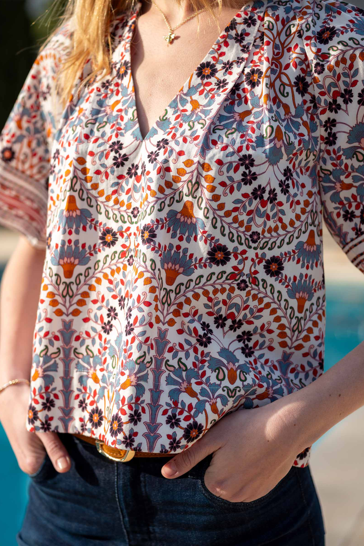 A close-up of a woman wearing a short-sleeve V-neck blouse with a colorful bohemian floral and paisley print in orange, blue, black, and white, paired with dark blue jeans.