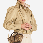 A model wearing a beige puffed-sleeve jacket, a cream lace skirt, and carrying a small brown and gold glittery handbag with a gold chain strap against a light gray studio background.
