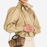 A model wearing a beige puffed-sleeve jacket, a cream lace skirt, and carrying a small brown and gold glittery handbag with a gold chain strap against a light gray studio background.