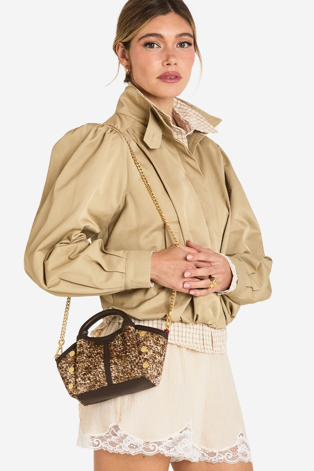 A model wearing a beige puffed-sleeve jacket, a cream lace skirt, and carrying a small brown and gold glittery handbag with a gold chain strap against a light gray studio background.