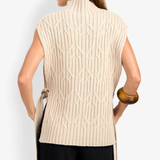 A woman shown from the back wearing a cream-colored sleeveless cable-knit vest with a mock neck and side tie, paired with black pants, against a light gray studio background.