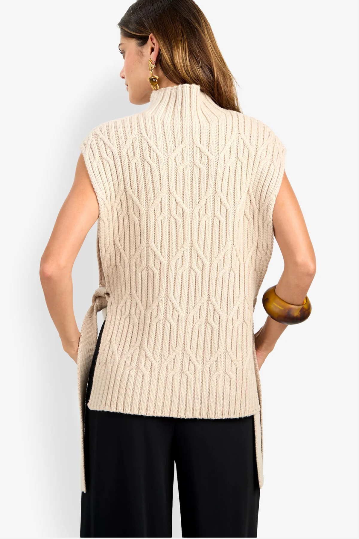 A woman shown from the back wearing a cream-colored sleeveless cable-knit vest with a mock neck and side tie, paired with black pants, against a light gray studio background.