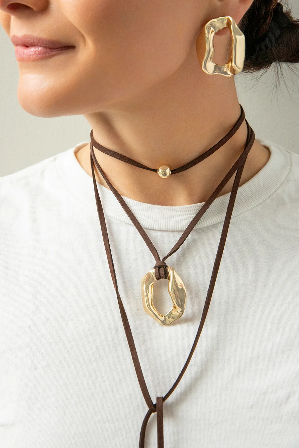 Close-up of a person wearing gold earrings and brown leather necklaces with gold pendants.