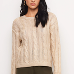 A woman modeling a beige cable-knit crewneck sweater paired with dark olive green pants, set against a plain white studio background.