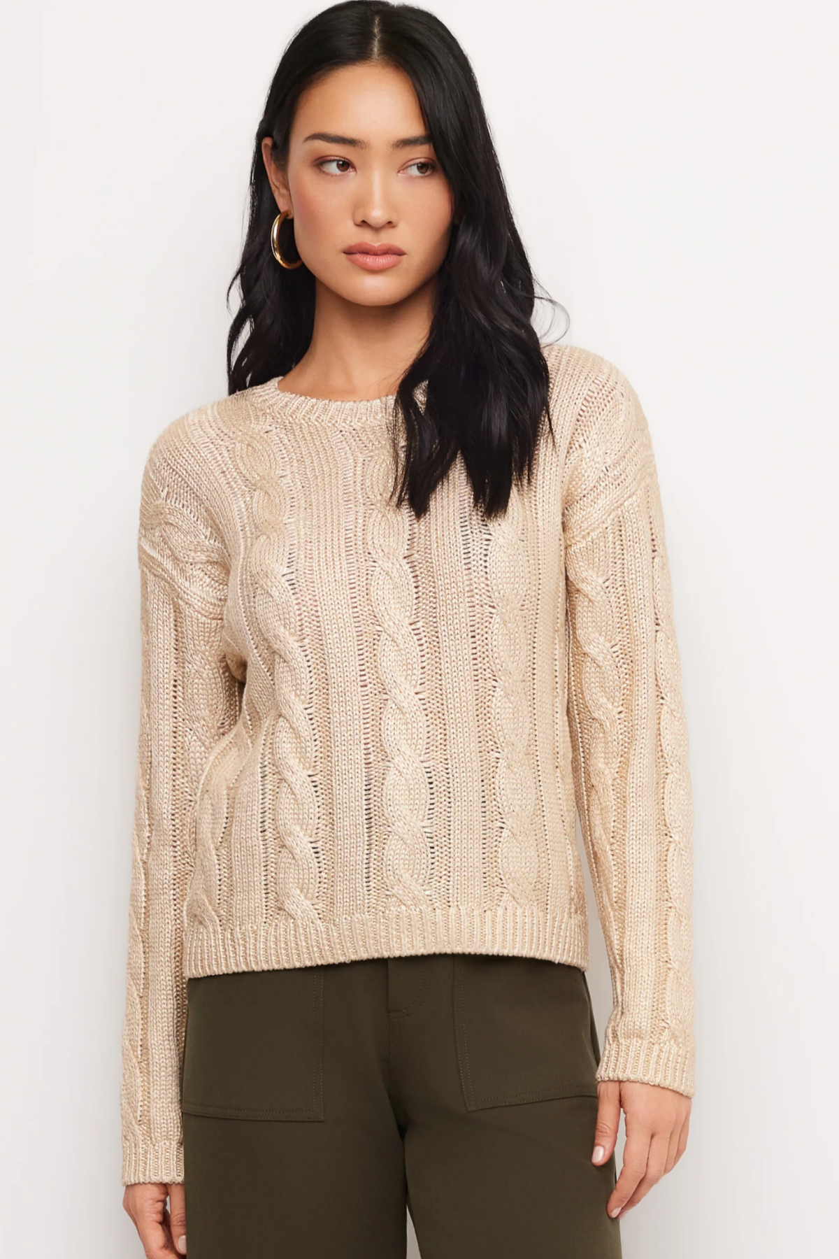 A woman modeling a beige cable-knit crewneck sweater paired with dark olive green pants, set against a plain white studio background.