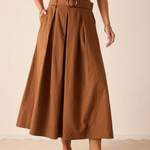 A model wearing a high-waisted, pleated brown midi skirt with a matching belt and round buckle, styled with a sage green top and dark brown leather loafers against a neutral beige background.