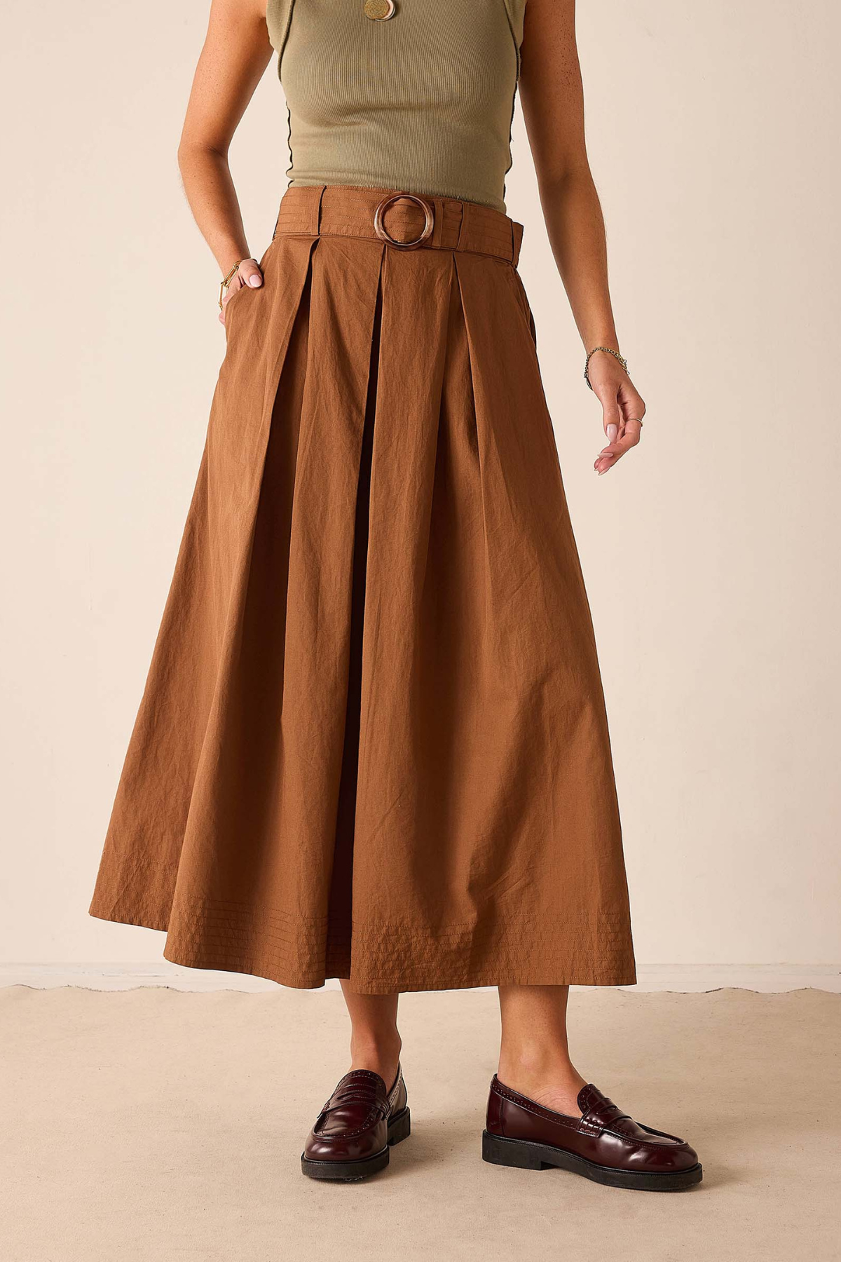 A model wearing a high-waisted, pleated brown midi skirt with a matching belt and round buckle, styled with a sage green top and dark brown leather loafers against a neutral beige background.