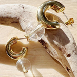 Gold-tone hoop earrings with clear acrylic beads, displayed on a piece of driftwood over a light wood surface.