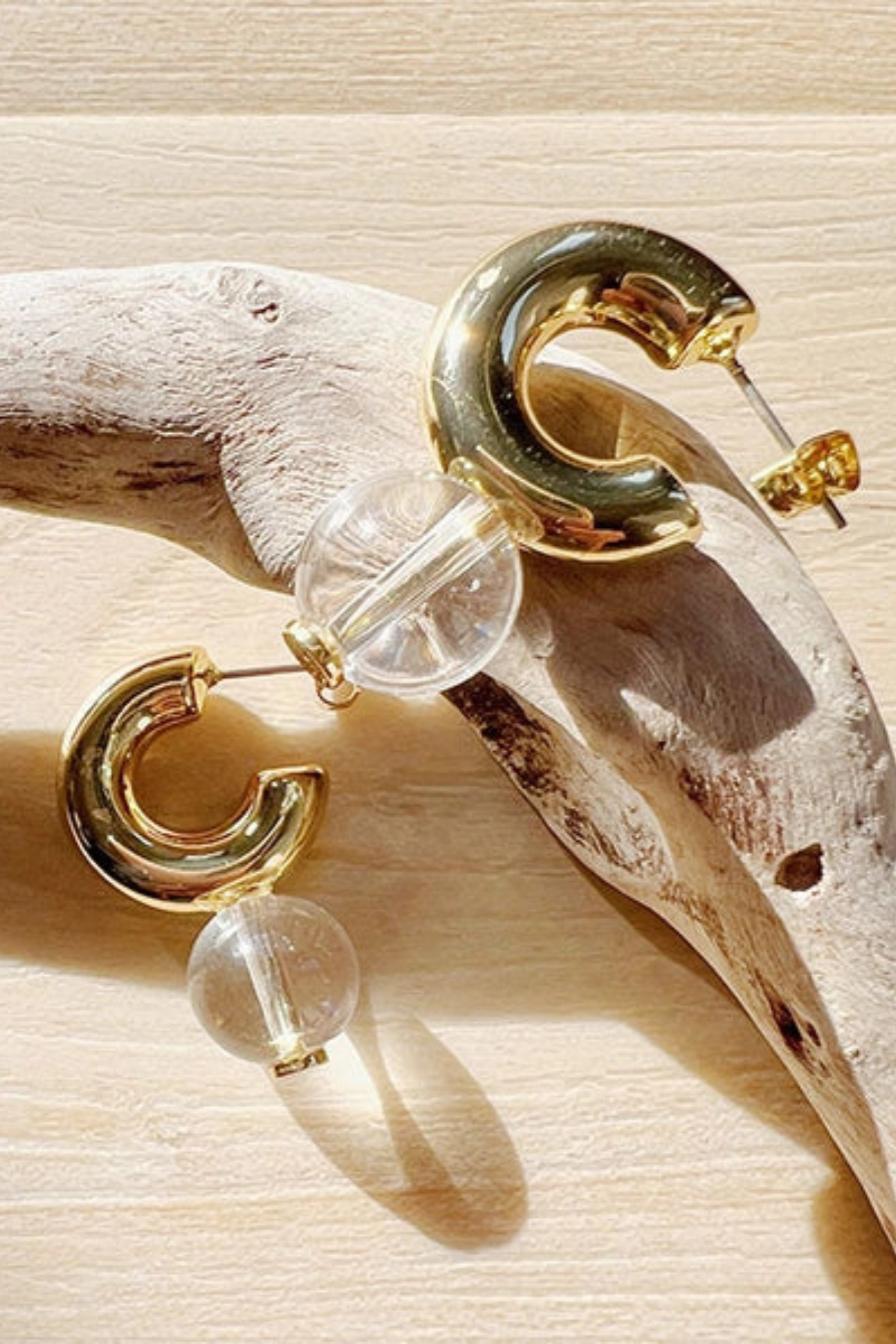 Gold-tone hoop earrings with clear acrylic beads, displayed on a piece of driftwood over a light wood surface.