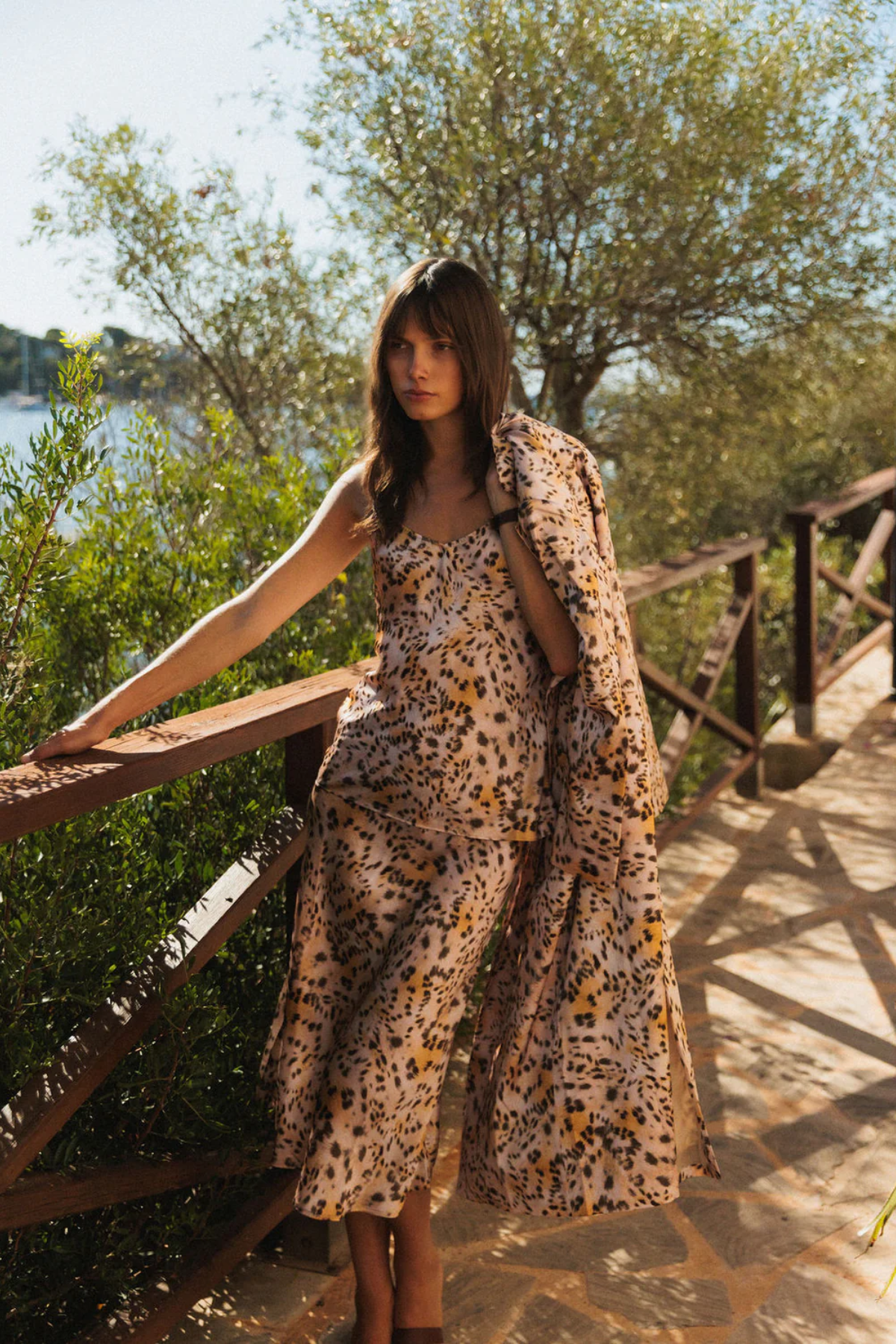 Woman in a leopard print dress standing on a wooden bridge with greenery and water in the background