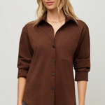 A woman wearing a brown button-up shirt with rolled sleeves and blue jeans, standing against a light gray studio background.
