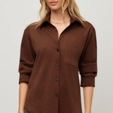 A woman wearing a brown button-up shirt with rolled sleeves and blue jeans, standing against a light gray studio background.