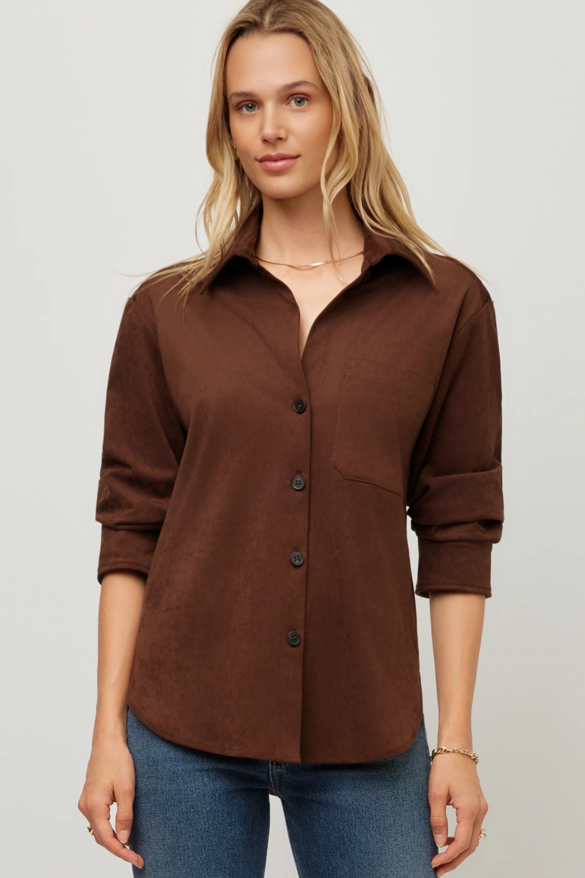 A woman wearing a brown button-up shirt with rolled sleeves and blue jeans, standing against a light gray studio background.