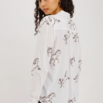 A woman wearing a white long-sleeve button-up shirt with an allover brown line-art print of running horses, paired with dark jeans, photographed against a plain light gray studio background.