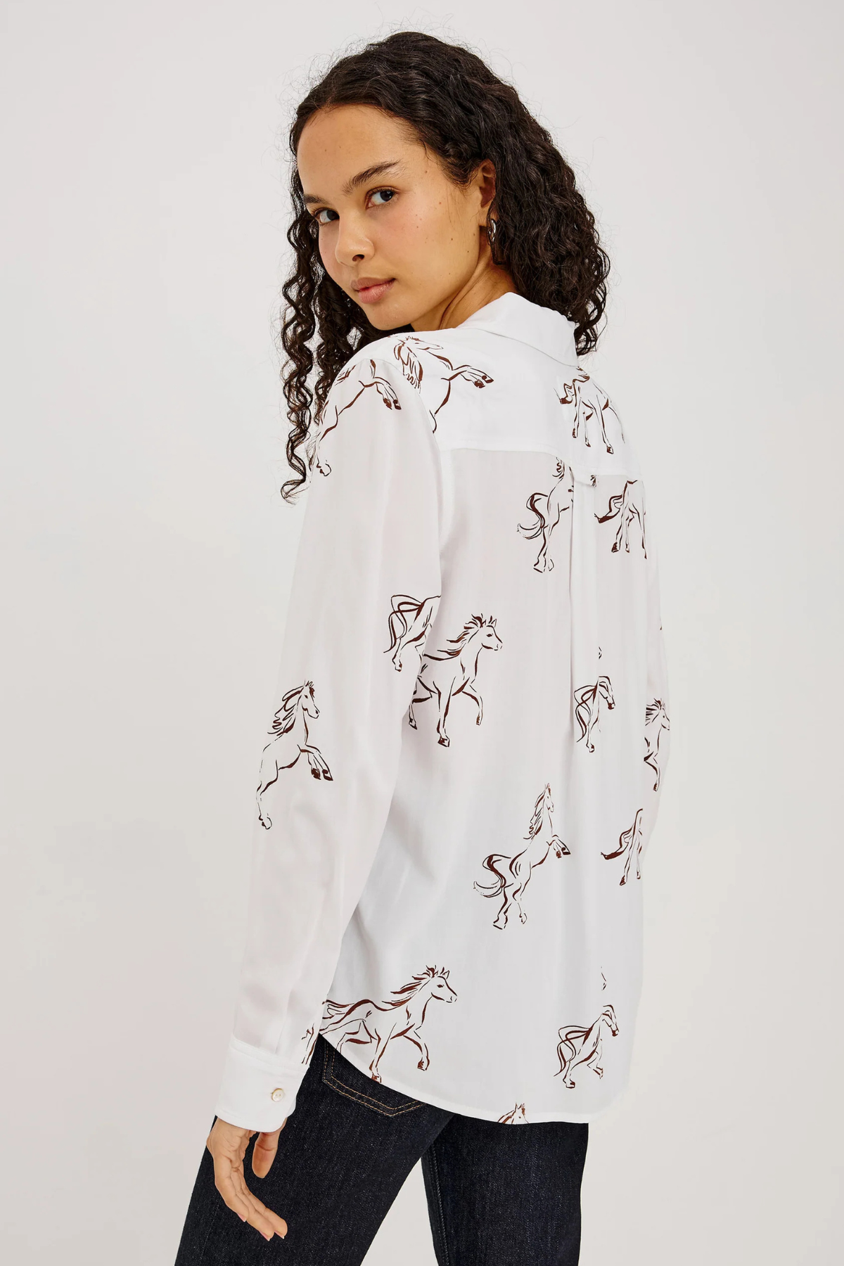 A woman wearing a white long-sleeve button-up shirt with an allover brown line-art print of running horses, paired with dark jeans, photographed against a plain light gray studio background.