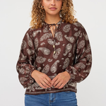 A woman with curly light brown hair wearing a brown long-sleeve blouse with a white paisley pattern and blue jeans, standing against a light gray studio background.