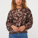 A woman with curly light brown hair wearing a brown long-sleeve blouse with a white paisley pattern and blue jeans, standing against a light gray studio background.