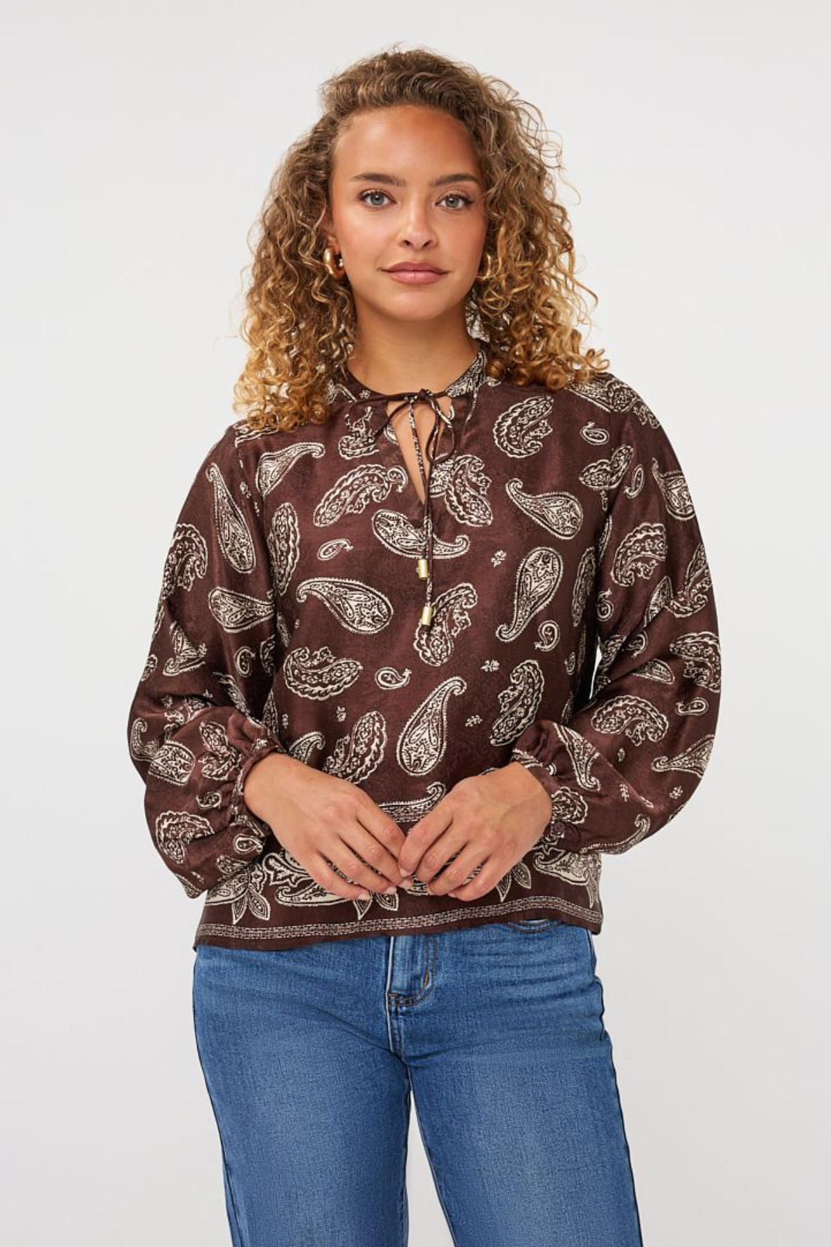 A woman with curly light brown hair wearing a brown long-sleeve blouse with a white paisley pattern and blue jeans, standing against a light gray studio background.