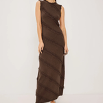 A woman modeling a sleeveless, high-neck, floor-length brown dress with diagonal textured fringe-like stripes, paired with black strappy heels, against a plain light gray studio background.