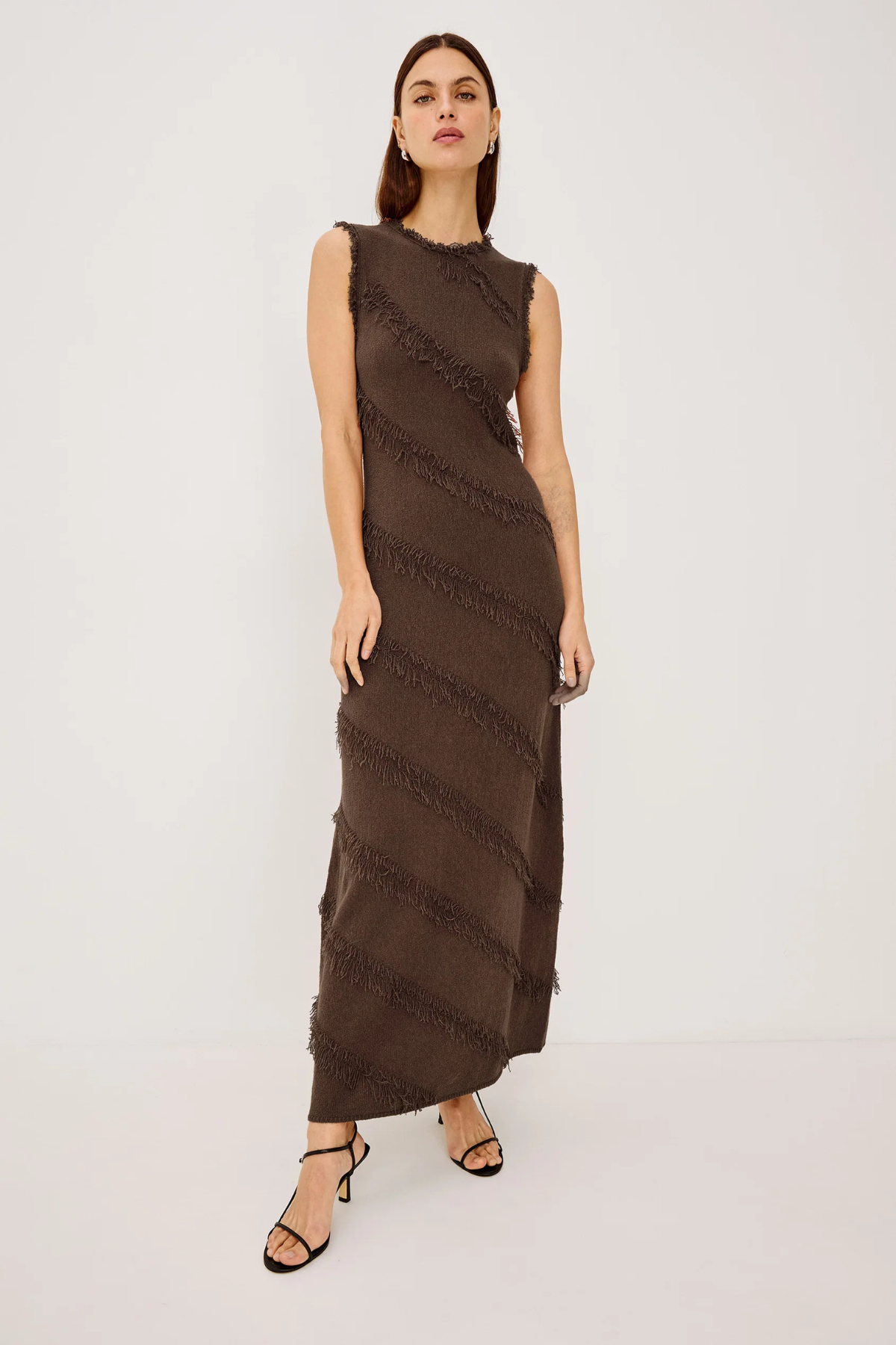 A woman modeling a sleeveless, high-neck, floor-length brown dress with diagonal textured fringe-like stripes, paired with black strappy heels, against a plain light gray studio background.