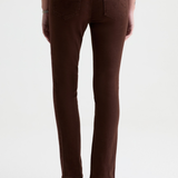 Rear view of a model wearing brown slim-fit jeans and black pointed-toe heels against a plain white studio background.
