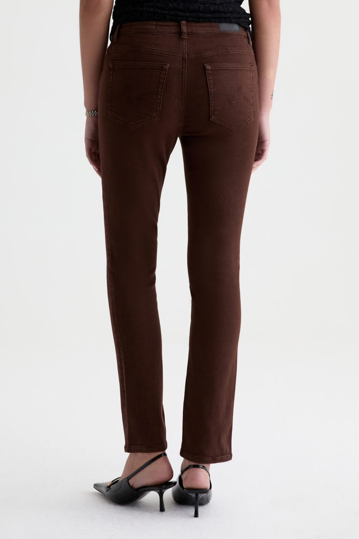 Rear view of a model wearing brown slim-fit jeans and black pointed-toe heels against a plain white studio background.