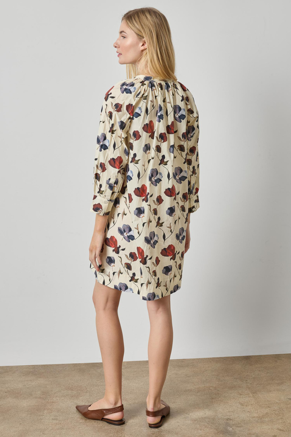A woman modeling a cream-colored mini dress with a red and blue floral pattern, shown from a side-back angle against a neutral studio background.