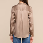 A woman viewed from the back wearing a long-sleeve satin blouse in a taupe-beige color, paired with blue jeans, against a solid beige background.