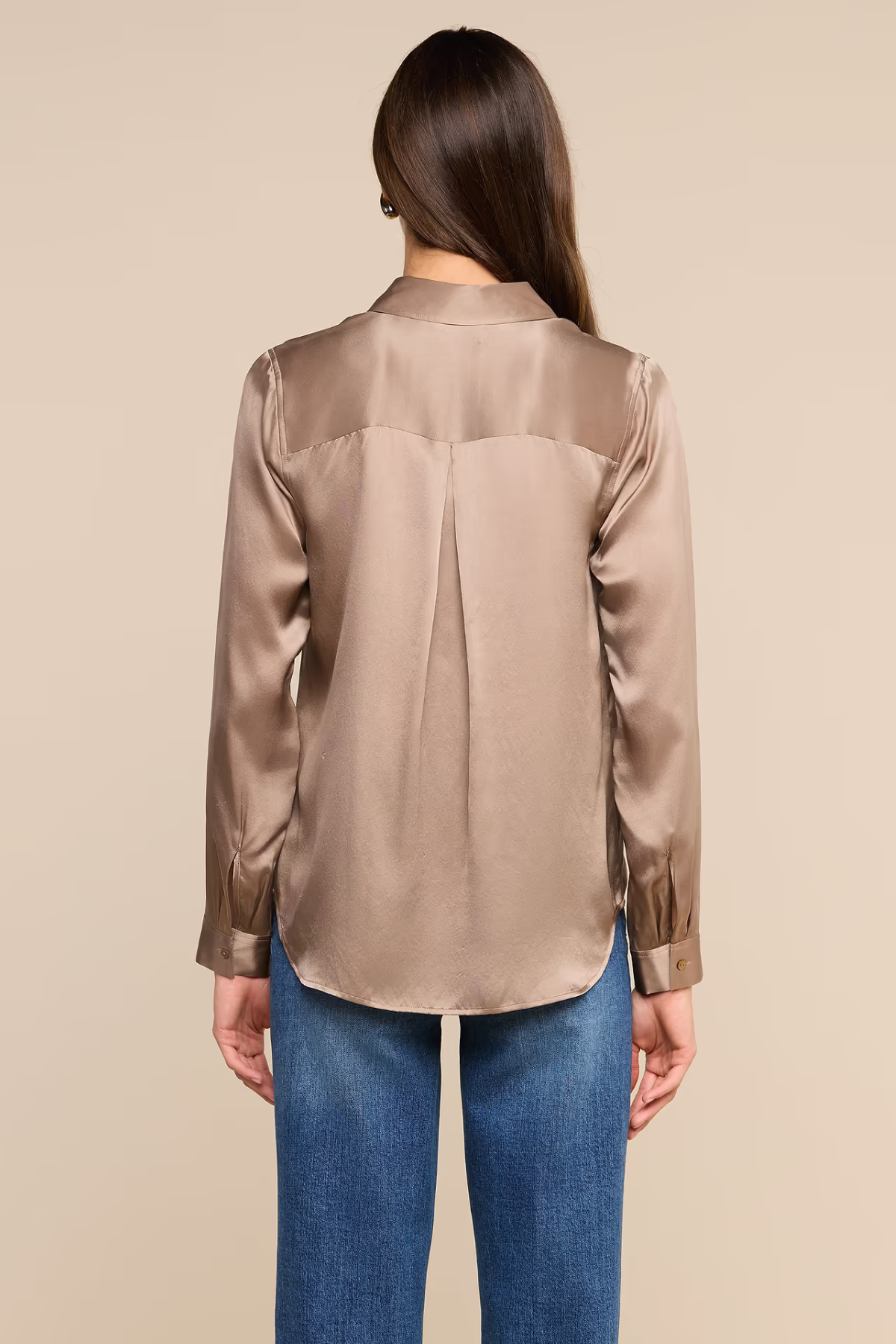 A woman viewed from the back wearing a long-sleeve satin blouse in a taupe-beige color, paired with blue jeans, against a solid beige background.