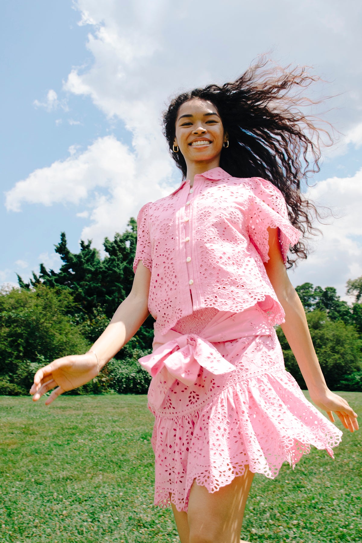 Woman in a pink outfit standing outdoors with trees and blue sky in the background