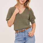 A woman modeling a short-sleeve ribbed knit top in olive green, paired with light blue jeans and a brown belt, against a plain light background.