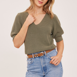 A woman modeling a short-sleeve ribbed knit top in olive green, paired with light blue jeans and a brown belt, against a plain light background.