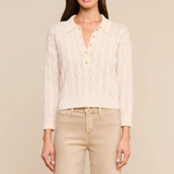 A woman modeling a cream cable-knit polo cardigan with gold buttons, paired with beige trousers, against a solid beige background.