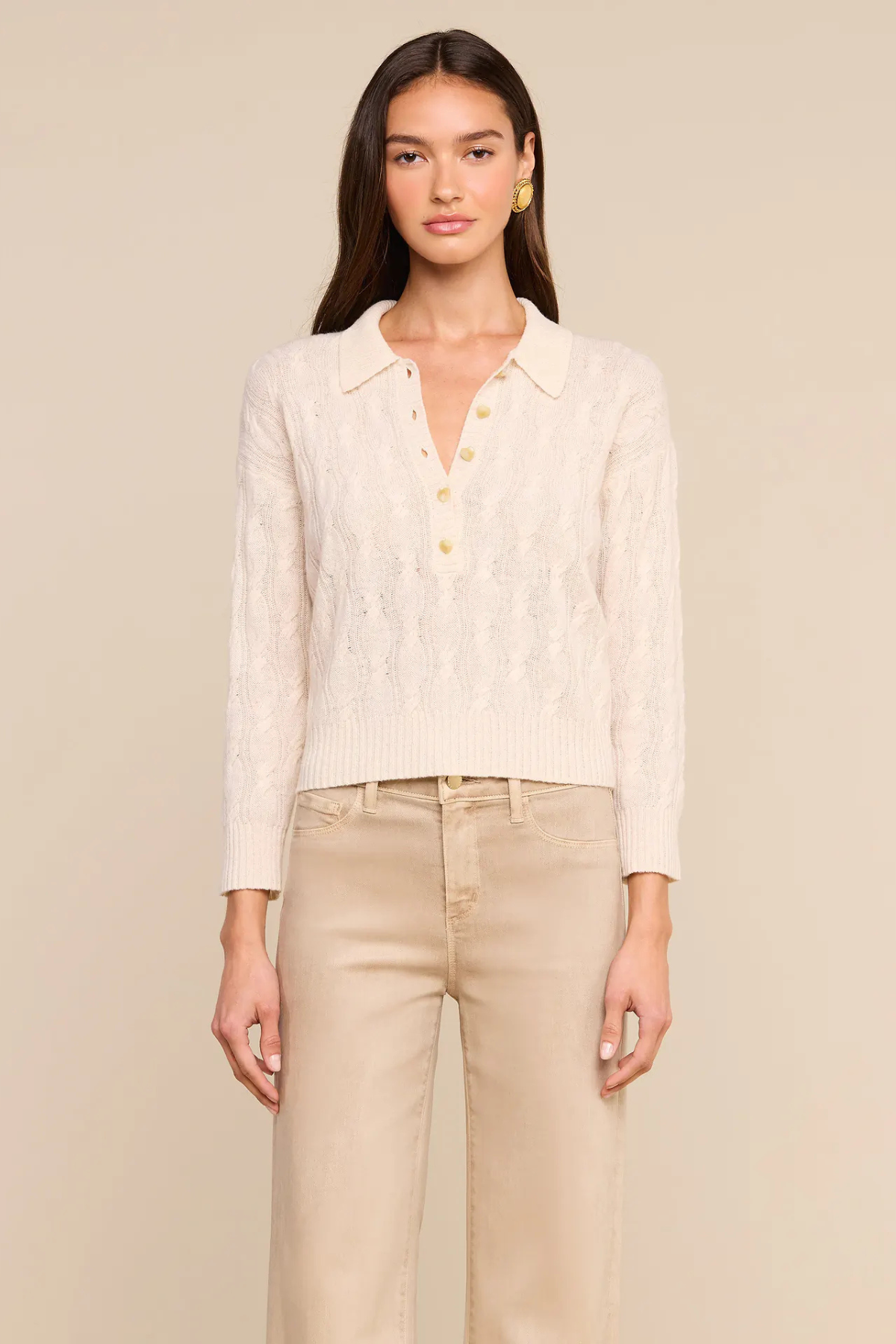 A woman modeling a cream cable-knit polo cardigan with gold buttons, paired with beige trousers, against a solid beige background.