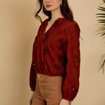 Woman wearing a red blouse with black patterns and brown pants against a beige background