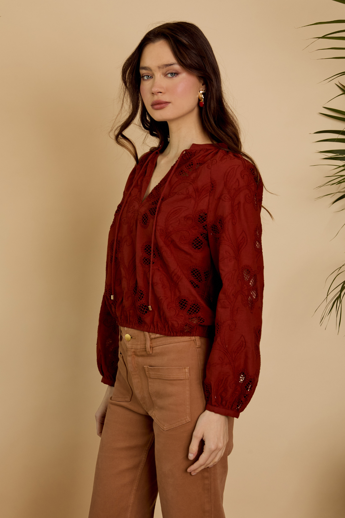 Woman wearing a red blouse with black patterns and brown pants against a beige background
