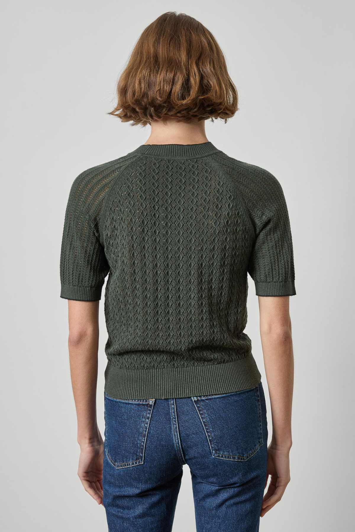 A person shown from the back wearing a dark olive green short-sleeve knit sweater with a textured cable pattern and blue jeans, against a plain light gray studio background.