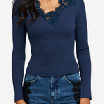 A woman wearing a navy blue long-sleeve top with dark lace detailing around the scoop neckline, paired with blue jeans featuring black lace side panels, against a light gray background.