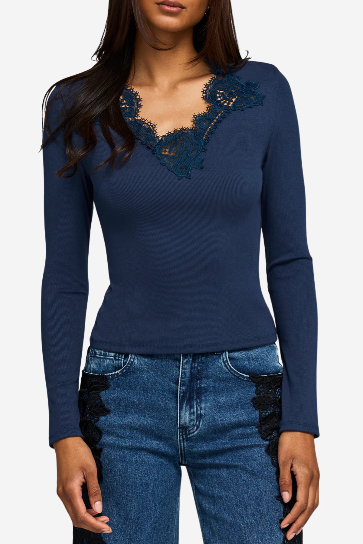 A woman wearing a navy blue long-sleeve top with dark lace detailing around the scoop neckline, paired with blue jeans featuring black lace side panels, against a light gray background.