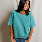 Person wearing a teal sweater and blue jeans against a plain background