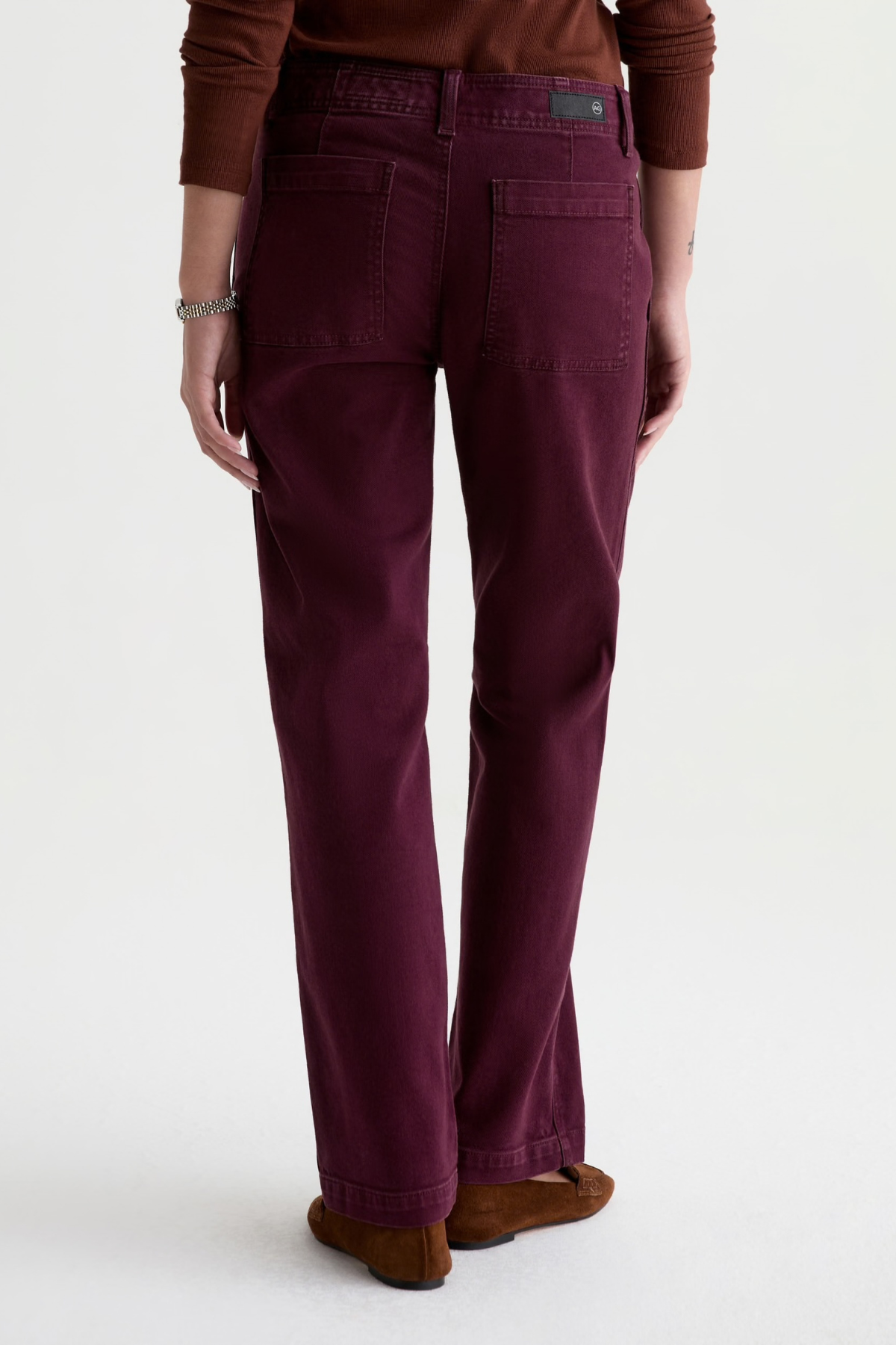 A person shown from the back wearing dark plum-colored straight-leg jeans, a brown long-sleeve top, and brown suede shoes against a plain white studio background.
