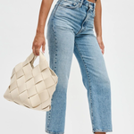 Person wearing light blue jeans and a black top, holding a woven beige bag on a white background