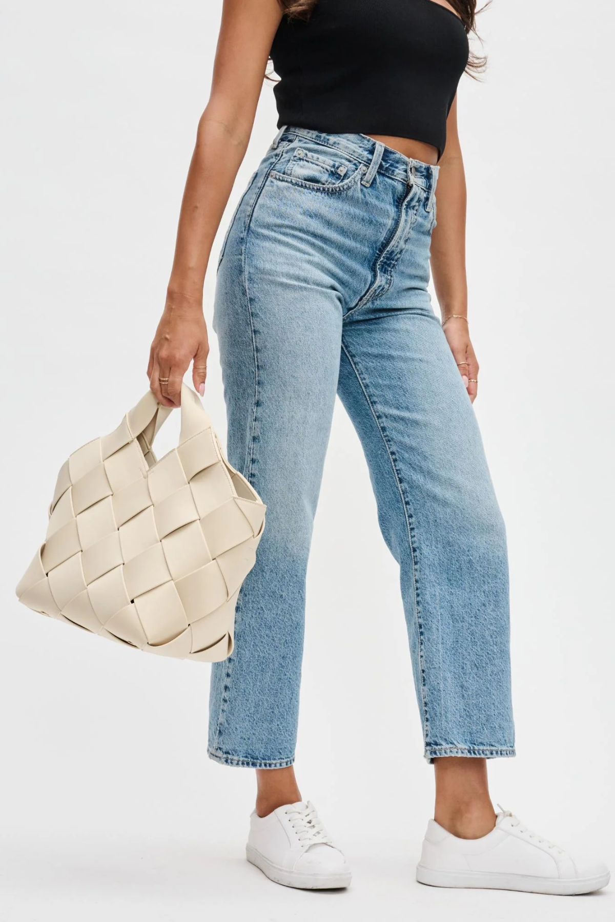 Person wearing light blue jeans and a black top, holding a woven beige bag on a white background