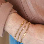 Three delicate gold chain bracelets with small letter charms worn stacked on a person's wrist, shown against a dusty rose top and blue denim background.